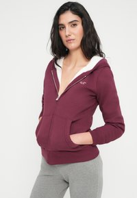 Μπορντό φούτερ με φερμουάρ, διαθέτει κουκούλα με λευκή επένδυση από φλις, μακριά μανίκια και μπροστινές τσέπες. Μαλακό ύφασμα με εφαρμοστή γραμμή.