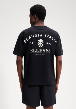 Mężczyzna stojący tyłem, ubrany w czarną koszulkę tenisową klubu Ellesse z białym napisem "Perugia Italia Dal 1959" oraz czarne spodenki na jednolitym tle.