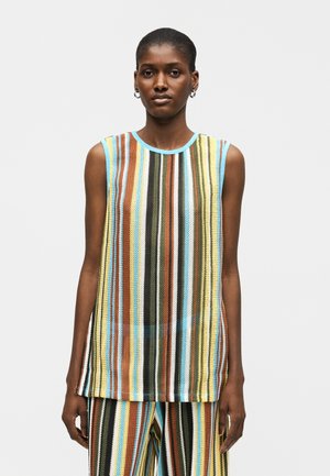 Γυναίκα που φοράει αμάνικη πλεκτή μπλούζα και αντίστοιχο παντελόνι με κάθετες ρίγες σε μπλε, καφέ, κίτρινο, μαύρο και λευκό, στέκεται μπροστά σε απλό φόντο.