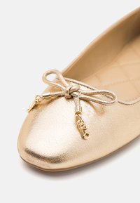 Ballerine plate dorée brillante avec un nœud noué sur le dessus, ornée de petits charms métalliques dorés aux extrémités du nœud et d'une doublure intérieure matelassée.