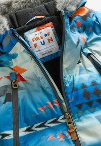 Chaqueta de abrigo colorida con patrones geométricos en azul, naranja y negro, con un interior forrado de forro polar y cremalleras de latón. Etiqueta visible.