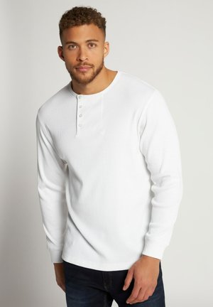 Camisa henley de manga larga blanca con textura acanalada, que presenta un cuello redondo y una tapeta con tres botones, combinada con jeans oscuros.