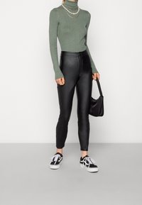 Personne portant un col roulé côtelé vert, un pantalon en cuir noir, des baskets noires et blanches, tenant un petit sac à main noir.