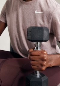 Osoba ubrana w różową koszulkę Nike i bordowe legginsy, trzymająca czarny heksagonalny hantel w jednej ręce, siedząca.