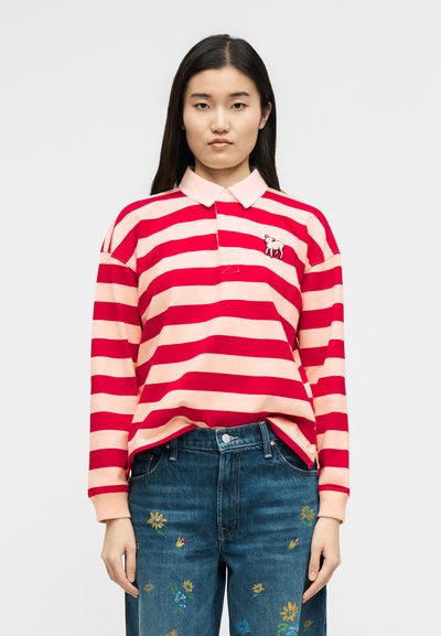 Γυναίκα που φοράει ριγέ ροζ και κόκκινο μπλουζάκι πόλο με μακριά μανίκια και ένα μικρό λογότυπο ζώου, συνδυασμένο με μπλε τζιν που έχει πολύχρωμη κεντημένη ανθοφορία.