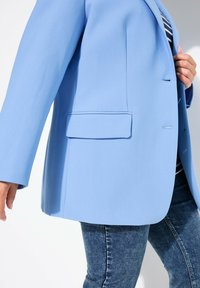 Blazer bleu clair en tissu structuré avec une poche unique, des revers pointus et une coupe décontractée, porté sur une chemise rayée et un jean.