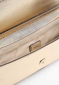 Kultainen tekonahkainen käsilaukku, jossa on teksturoitu sisustus, vetoketjullinen tasku ja merkkilogo-tarra. Varustettu napilla suljettavalla kannella ja sileällä vuorilla.