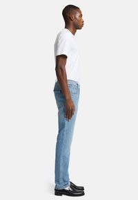 Hellblaue Denim-Jeans, Slim Fit, mit normalem mittelhohem Bund und fünf Taschen. Kombiniert mit einem weißen T-Shirt mit kurzen Ärmeln und schwarzen Schuhen.