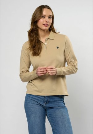 Beige pitkähihaista polo-paitaa, jossa on kaulus, ja vasemmassa rinnassa pieni musta emblem sekä hienovarainen tekstuuri. Yhdistettynä sinisiin farkkuihin.