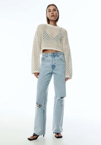 Knit-Crop-Pullover in Creme mit offenem Maschenmuster, kombiniert mit hellblauen, verwaschenen Jeans, die an den Knien und Knöcheln Risse aufweisen.
