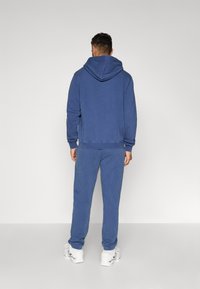 Μπλε βαμβακερή φόρμα με κουκούλα και κορδόνι, σε συνδυασμό με μπλε παντελόνι φόρμας. Και τα δύο έχουν χαλαρή εφαρμογή και ριγέ μανσέτες.