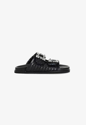 Black woven slide sandals feature dual straps adorned with rectangular, sparkly rhinestone buckles and a flat, textured rubber sole.