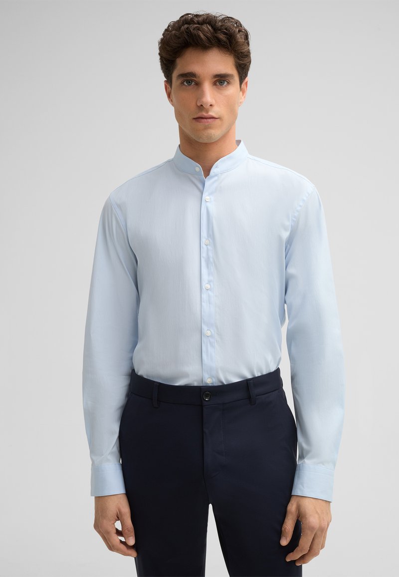 Lichtblauw overhemd met lange mouwen en een bandkraag, met een soepele textuur en slim fit. Draagt bij donkere marineblauwe broek.