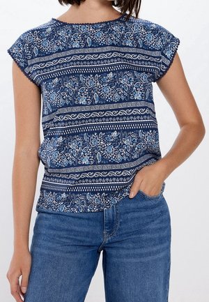 Kobieta ubrana w niebieską bluzkę z krótkim rękawem w kwiatowy wzór, zestawioną z niebieskimi dżinsami, ręka swobodnie w kieszeni na jednolitym tle.