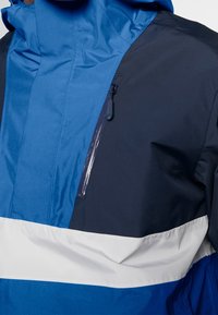 Giacca resistente all'acqua caratterizzata da una combinazione di blu e navy, con una striscia orizzontale bianca e una tasca sul petto con zip.
