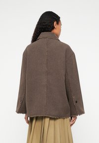 Bruine, oversized jas gemaakt van textuurstof, met een notch-kraag, zichtbare stiksels en een rafelige zoom. Knopdetail op de mouw.