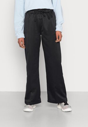Personne portant un pantalon noir à jambes larges avec une taille à cordon, un haut bleu clair à manches longues et des baskets blanches et grises, debout sur un sol gris.