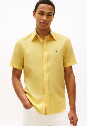 Junger Mann, der ein kurzärmeliges gelbes Hemd mit kleinen Logo auf der Brust und weiße Hose trägt, steht mit einer Hand in der Tasche.