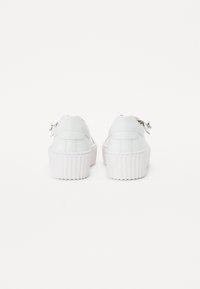 Hvide platformsneakers med teksturerede gummisåler, glat læderoverdel og dekorative metalliske detaljer på hælen.
