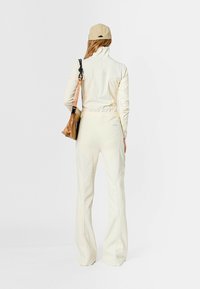 Conjunto deportivo de color beige claro que incluye una chaqueta con cremallera de cuello alto y pantalones acampanados con costuras curvas. Una gorra color caqui y un bolso beige completan el look.