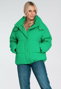 Zielona kurtka puchowa z wysokim kołnierzem, z zapięciem na napy i bocznymi kieszeniami. Teksturowane wykończenie, luźny krój. Nosić z dżinsowymi spodniami.