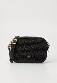 Μαύρη τσάντα ώμου με υφή κυματιστού μοτίβου, χρυσό λογότυπο cK, ρυθμιζόμενο λουρί και επάνω κλείσιμο με φερμουάρ σε λευκό φόντο.