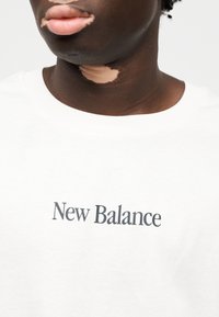 Λευκό βαμβακερό μπλουζάκι με το λογότυπο "New Balance" σε έντονη, σκουρόχρωμη γραμματοσειρά στο μπροστινό μέρος. Το ύφασμα φαίνεται λείο και απαλό.