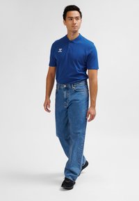 Niebieska koszulka polo z logo, zestawiona z luźnymi, jasnoniebieskimi dżinsami i czarnymi tenisówkami, na jednolitym szarym tle.