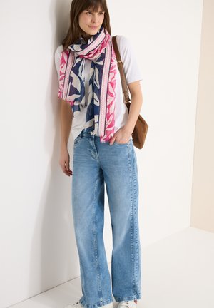 Femme appuyée contre un mur, portant un t-shirt blanc, un jean bleu à jambes larges, une écharpe rose et marine à motifs, et un sac à bandoulière marron.