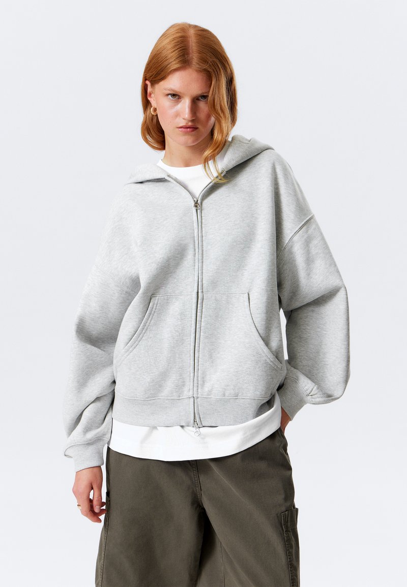 Grå zip-up hettegenser med stor, justerbar hette, senkede skuldre, frontlommer og ribbestrikkede mansjetter, lagd over en hvit skjorte.