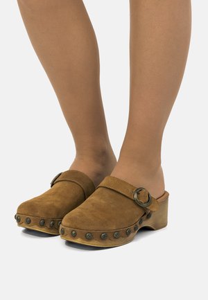 Sabots en daim marron avec des clous ronds en métal le long de la semelle et une bride décorative à boucle portés sur des pieds avec des bas couleur sable.