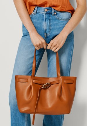 Ženska, oblečena v oranžni vrhnji del in modre kavbojke, drži bež usnjeno torbo tote z zlatim zaponkom na belem ozadju.