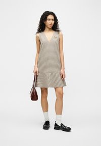 Γυναίκα με σγουρά μαλλιά που φοράει αμάνικο φόρεμα με καρό σχέδιο, λευκές κάλτσες, μαύρα λουστρινένια παπούτσια και κρατά καφέ τσάντα σε απλό φόντο.