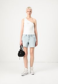 Hvit topp med én skulder som har et sommerfugldesign, sammen med lyseblå denimshorts og en svart veske med blomsterdetaljer. Hvite høye sneakers.