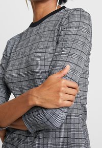 Top ajusté noir et blanc avec un motif à carreaux, présentant un col rond et des manches trois-quarts avec une bordure noire unie. Texture lisse.
