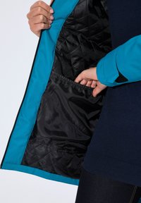 Blauwe jack met een zwart gevoerd interieur; toont een zak aan de binnenkant en een close-up van een hand die de rits vasthoudt.