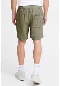 Olive green shorts with a textured pattern, elastic waistband, and a single back pocket. Styled with white athletic shoes and socks.