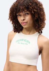 Νέα γυναίκα με σγουρά μαλλιά φοράει λευκό crop top Calvin Klein με το κείμενο «New York City» σε απλό φόντο.