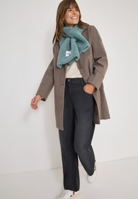 Brun uldfrakke over en cremet sweater, parret med mørke jeans og hvide sneakers. Et teal fleece-scarf er draperet rundt om halsen.