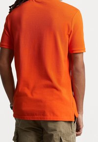 Pomarańczowa koszulka polo z teksturowanej tkaniny; z krótkimi rękawami, prostym dołem z bocznymi rozporkami i gładkim wzornictwem bez ozdób.