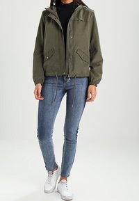 Chaqueta ligera con capucha de color verde oliva, con cierre de cremallera en la parte delantera, dobladillo ajustable con cordón, bolsillos laterales, llevada sobre una camiseta negra y jeans azules.