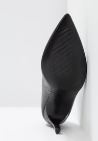 Escarpin noir en cuir à talon stiletto avec un bout pointu, montrant une semelle côtelée et une texture lisse sur le côté. Positionné sur un fond blanc.