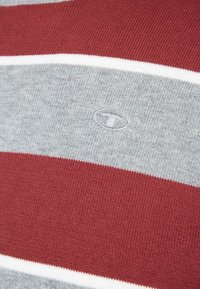 Nærbillede af strikket stof med vandrette striber i maroon, grå og hvid, der har et lille broderet logo i midten.