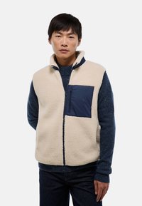 Gilet en polaire crème avec une poche poitrine zippée marine. Il présente un col montant et des accents d'épaules contrastants en marine. Texture lisse.