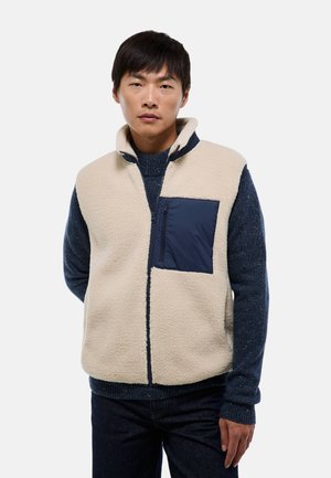 Gilet en polaire crème avec une poche poitrine zippée marine. Il présente un col montant et des accents d'épaules contrastants en marine. Texture lisse.