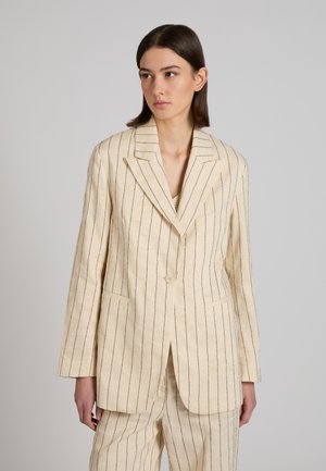 Jeune femme portant un costume couleur crème avec de fines rayures verticales noires, debout devant un fond gris clair uni.