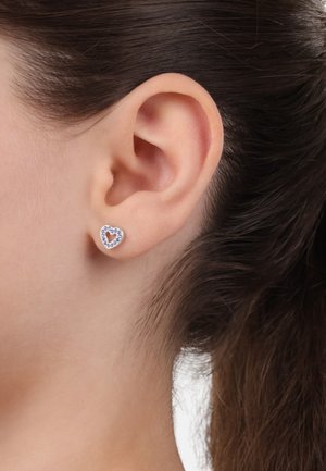 Orecchini a bottone a forma di cuore in argento, con un centro di gemma viola e un alone di piccole pietre chiare, mostrati all'orecchio di un modello.