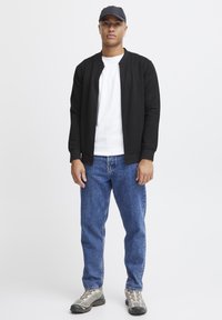 Cazadora bomber negra con textura sobre una camiseta blanca, combinada con pantalones vaqueros azules y zapatillas grises. El conjunto se complementa con una gorra oscura.