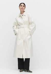 Biała trenchcoat z paskiem w talii, dużym kołnierzem i szerokimi rękawami. Modelka nosi czarne spodnie i buty w wzór węża.