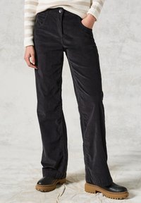 Pantalons en velours côtelé noirs avec une coupe droite, poches avant et fermeture par bouton. Associés à des chaussures plates noires.